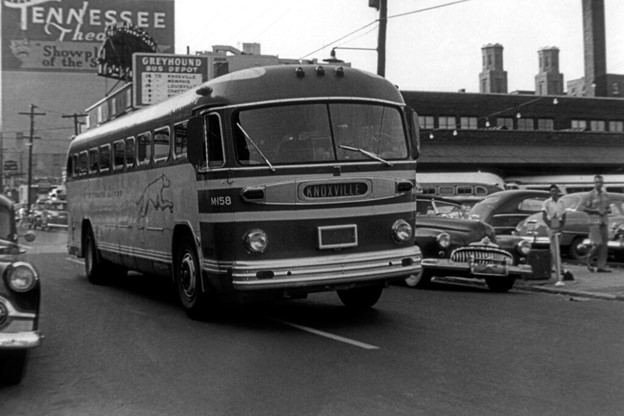 A historical black and white photo capturing a busy urban street scene featuring a Greyhound bus prominently in the foreground with the destination "KNOXVILLE" displayed above the windshield. The bus has a sleek design indicative of mid-20th-century automotive aesthetics, with a Greyhound dog logo and the fleet number M158 visible. The street is bustling with vintage cars parked along the curb, and pedestrians are seen in the background. In the upper left corner, the marquee of the Tennessee Theatre advertises "Show of the South." Architectural details of buildings and industrial structures, including smokestacks, are visible in the distant background, contributing to the mid-century American city atmosphere.