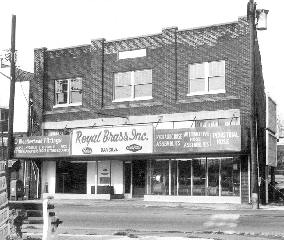 A black and white photo of a two-story brick commercial building. The ground level features large glass windows and a central glass door, typical of retail storefronts. Above the entrance, a prominent sign reads "Royal Brass Inc." with the company's offerings listed: "Hydraulic Hose Assemblies, Automotive Hose Assemblies, Industrial Hose." To the left, smaller signs advertise "Weatherhead Fittings, Brass, Steel, Stainless, Hydraulic Tube Fittings, Hose Clamps." The upper story of the building has six symmetrically placed double-hung windows. The architecture suggests a mid-20th-century style, common in small town or urban business districts. There are no visible people, and the street in front is empty, giving the scene a quiet, perhaps early morning or weekend feel.